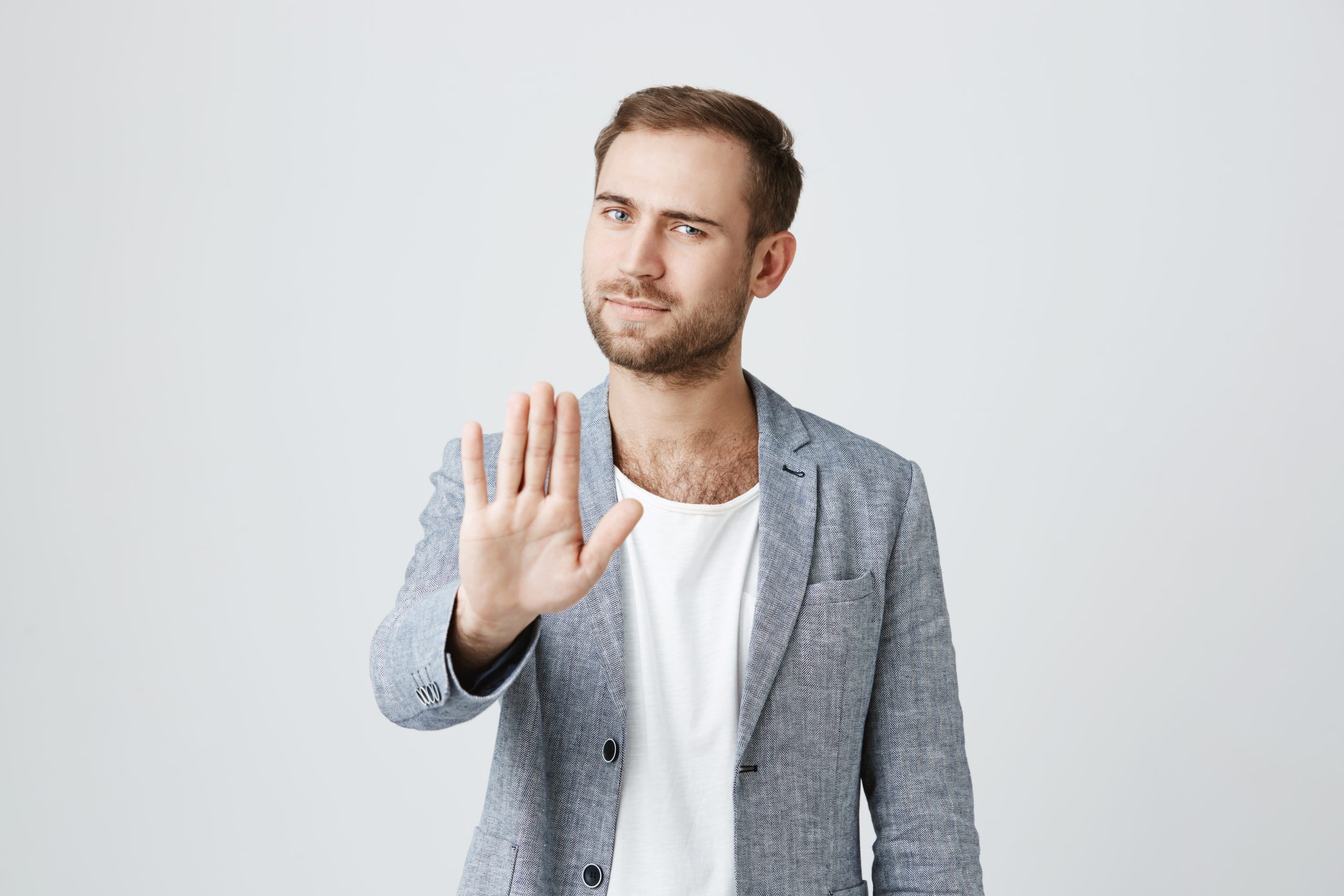 Angry bearded male model gestures indoors, shows stop sign with palm, refuses to do something, isolated against gray studio background. Serious bearded irritated man shows stop gesture indoors