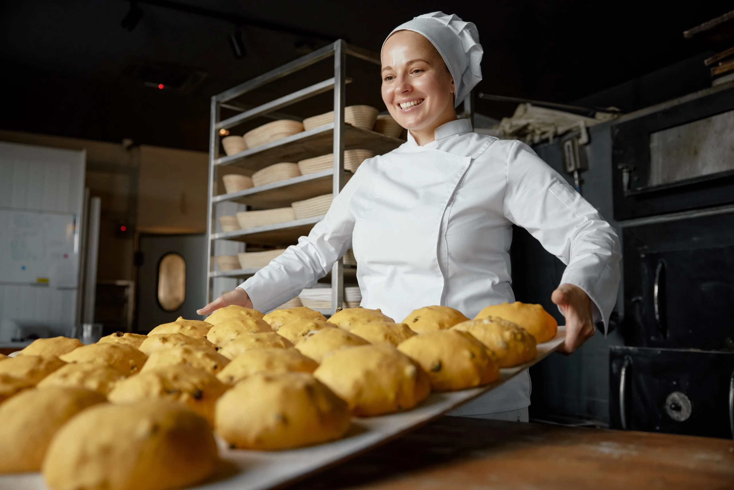 Uśmiechnięta piekarka w białym fartuchu niosąca tacę z upieczonym pieczywem w zakładzie produkcyjnym