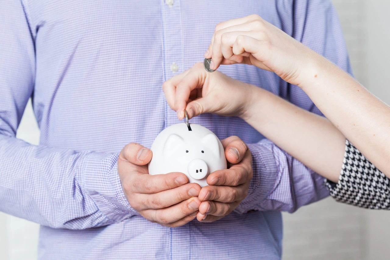Ręce mężczyzny trzymające skarbonkę w kształcie świnki, do której kobieta wkłada monetę – symbol oszczędzania na lokacie