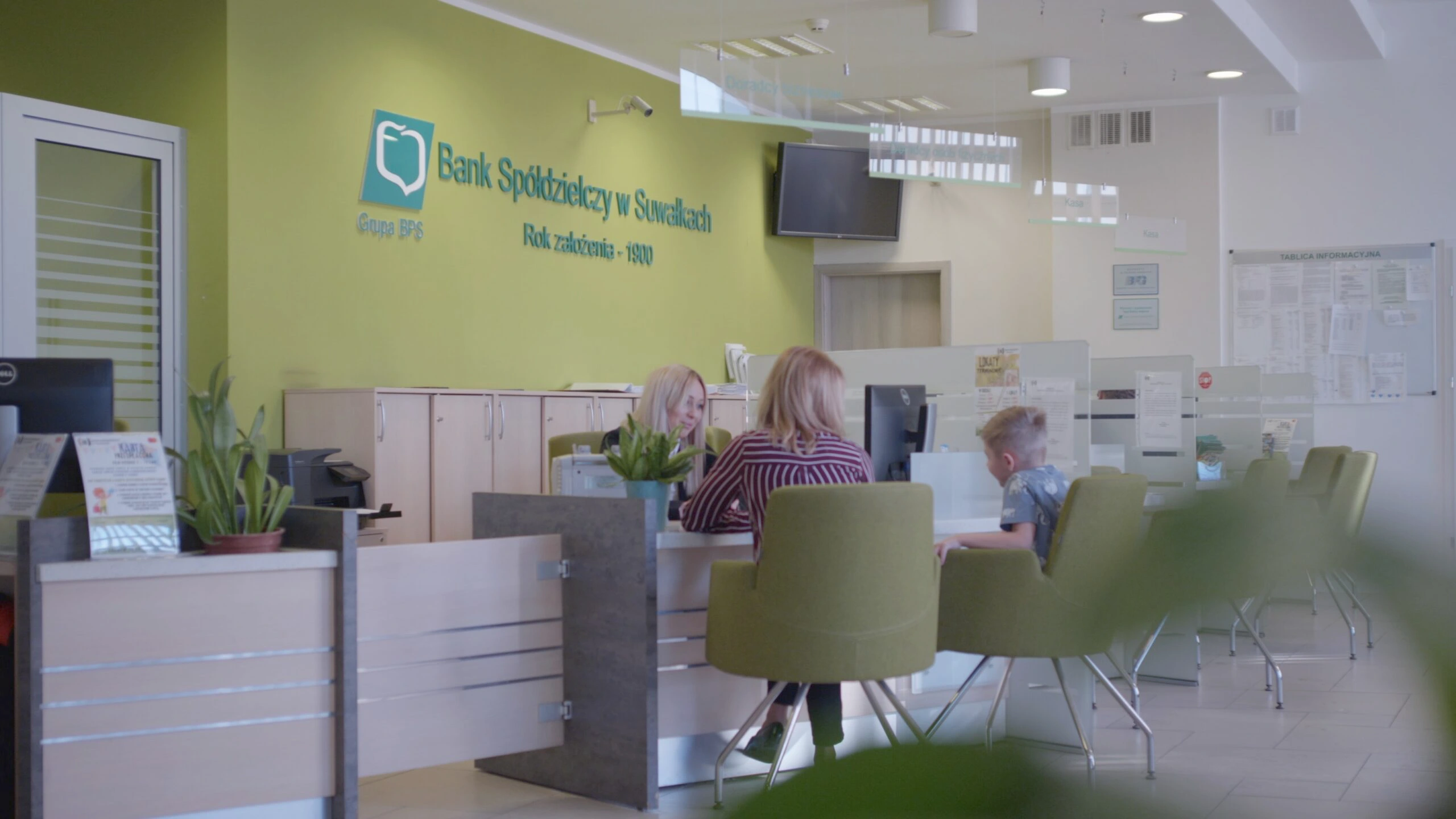 Stanowisko obsługi klienta w placówce Banku Spółdzielczego w Suwałkach z widoczną doradczynią, klientką i dzieckiem siedzącymi przy biurku w nowoczesnym wnętrzu