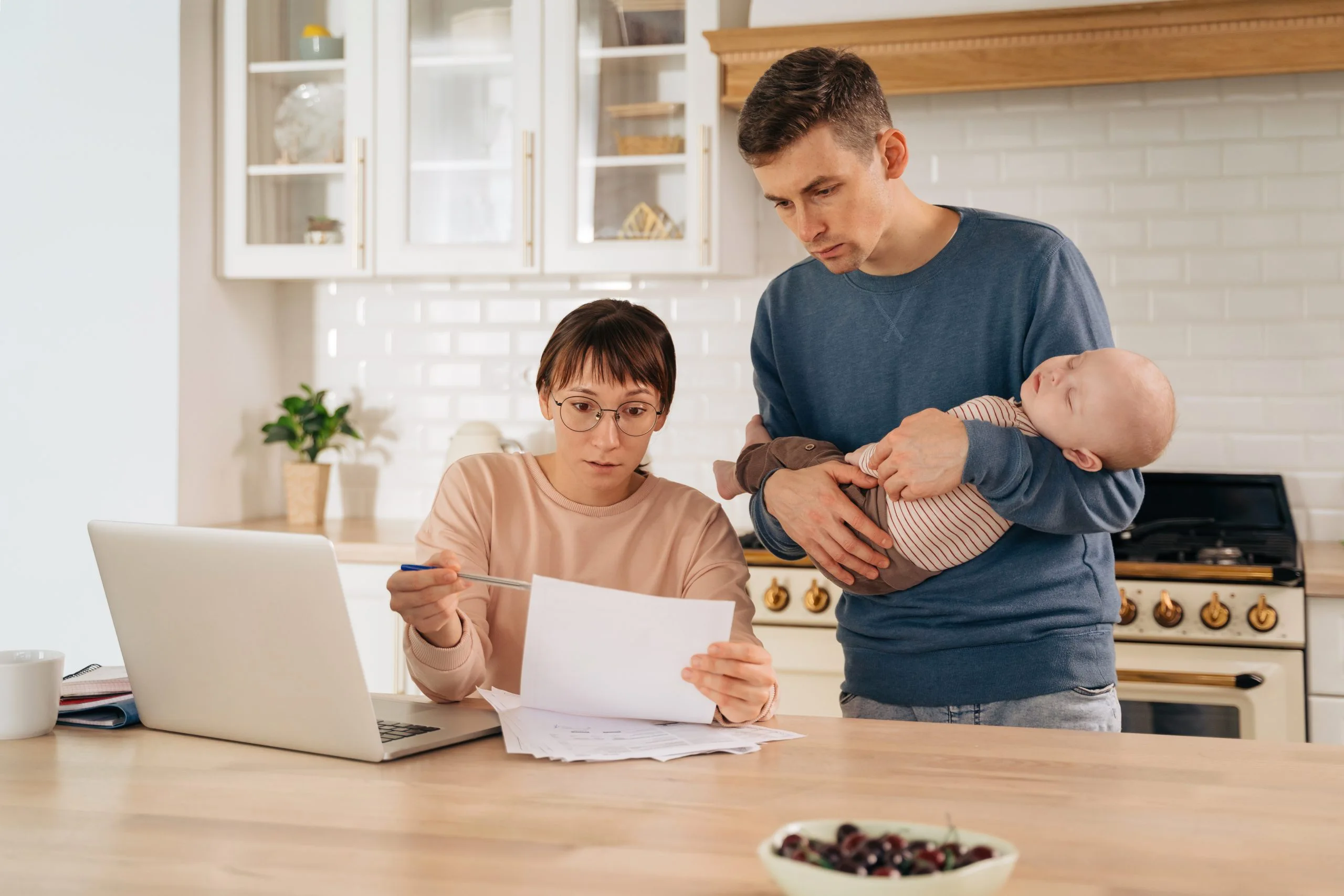 Młoda para z niemowlęciem analizująca dokumenty przy kuchennym stole, obok laptopa i filiżanki.