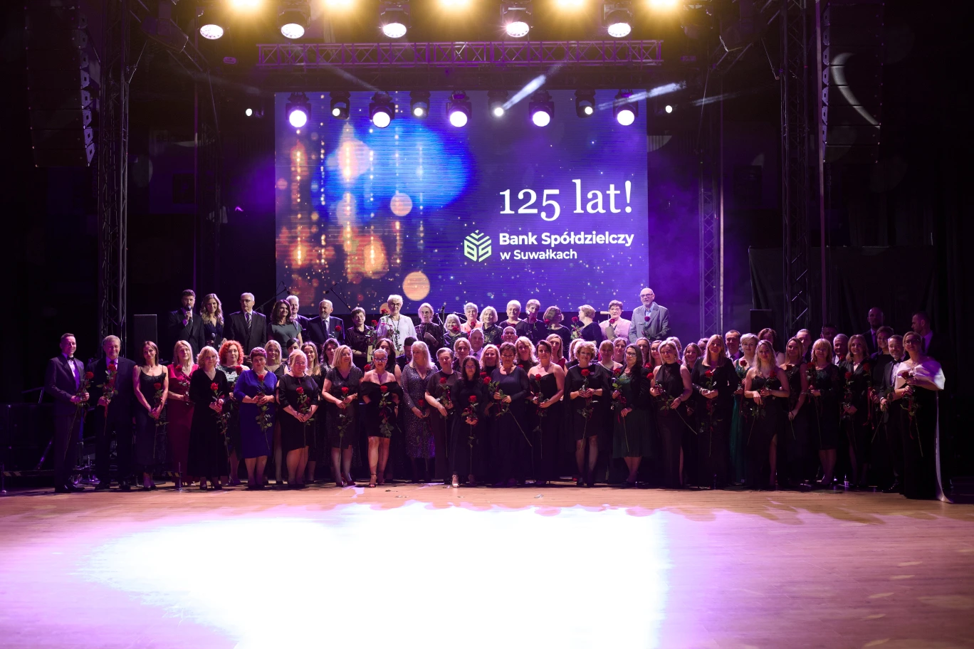Grupa pracowników Banku Spółdzielczego w Suwałkach pozująca do pamiątkowego zdjęcia na scenie podczas gali 125-lecia, trzymających w dłoniach róże jako symbol podziękowania.