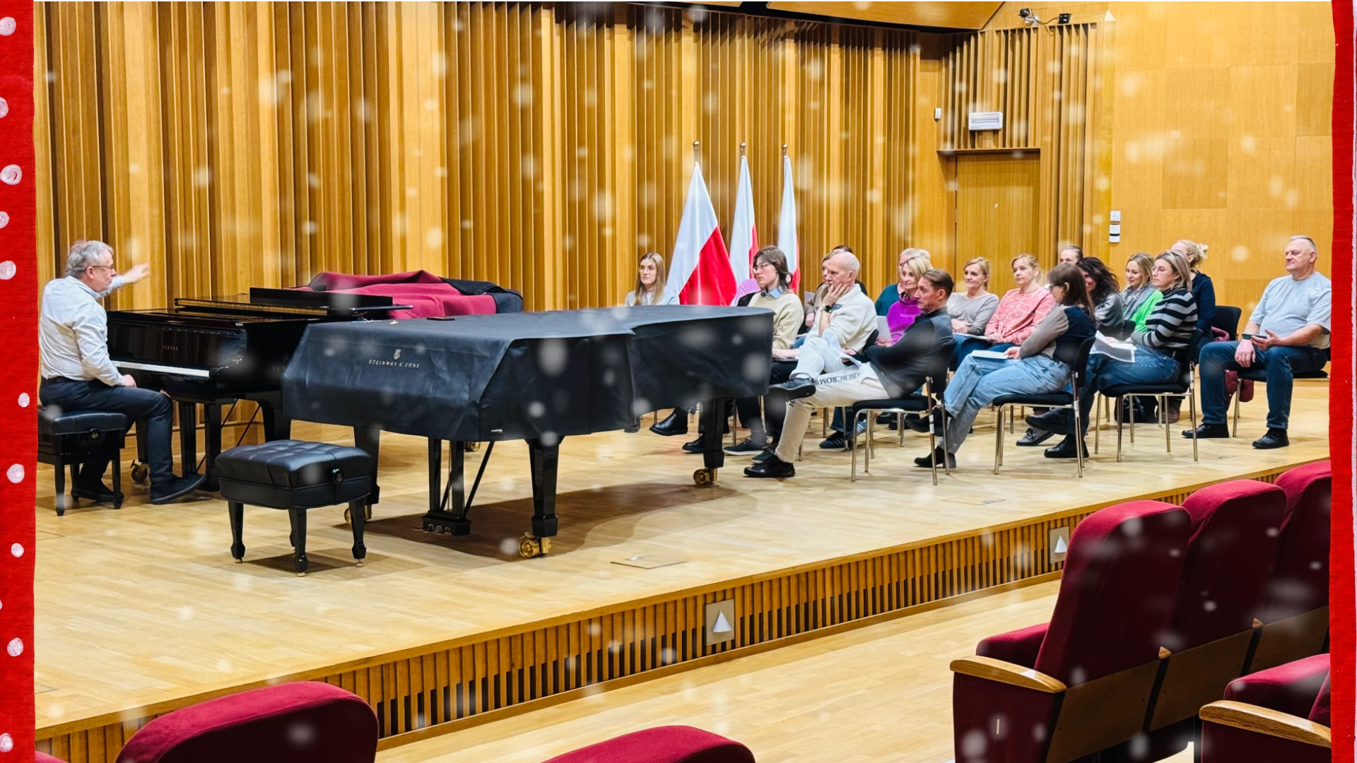 Grupa osób mężczyzn i kobiet siedzi na sali koncertowej podczas próby kolędowej, prowadzoną przy fortepianie przez muzyka.